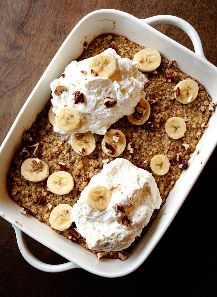 Overhead view of baked banana oatmeal topped with whipped cream, fresh banana slices, and chopped pecans in a white baking dish