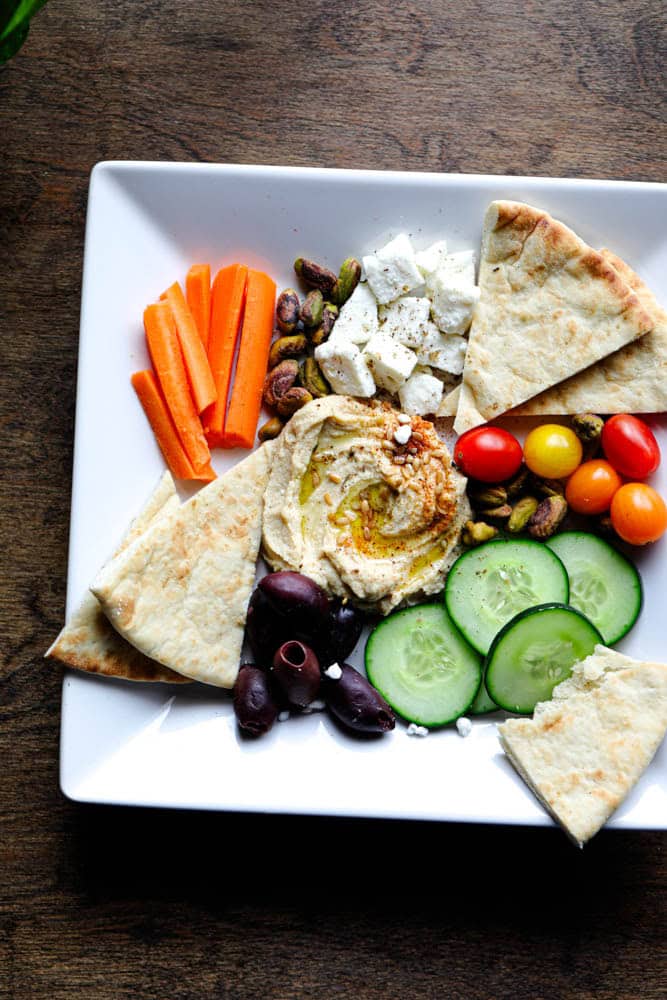 Overhead view of a Mediterranean hummus snack plate with vegetables, feta cheese, olives, pita bread, and pistachios